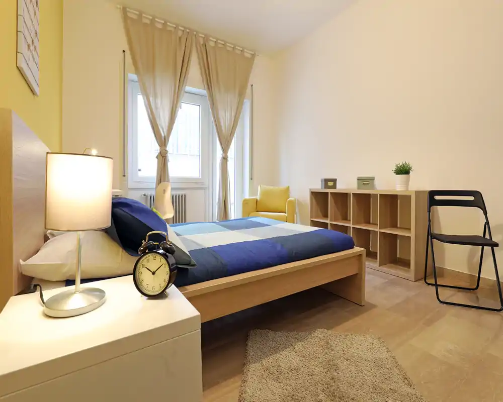 Chambre lumineuse et moderne avec un lit double en bois habillé de linge bleu, table de chevet avec lampe et réveil, tapis confortable et petite étagère; la lumière naturelle de la fenêtre et les accents chaleureux offrent une ambiance accueillante.