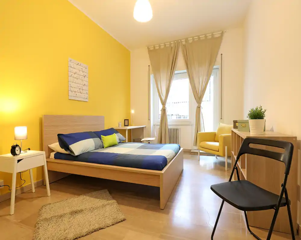 Chambre moderne et lumineuse avec lit double devant un mur jaune chaleureux, textiles doux et fauteuil près de la fenêtre — lumineuse et accueillante pour une location.