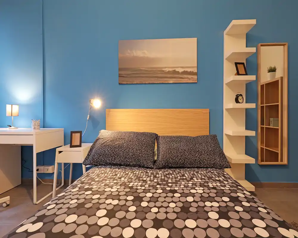 Bright, well-styled bedroom with a double bed centered against a blue accent wall, patterned bedding, bedside lamps and a small desk — cozy and modern.
