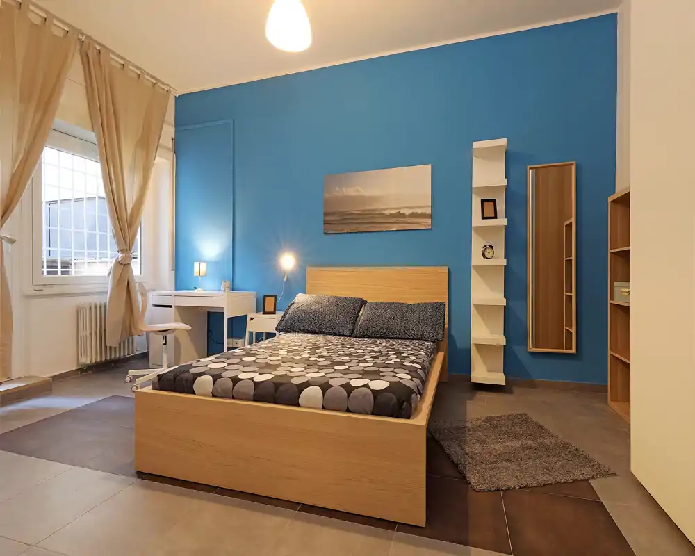 Bright, modern bedroom with a wooden double bed against a bold blue accent wall, a small desk by the window and warm layered lighting creating a cozy atmosphere.
