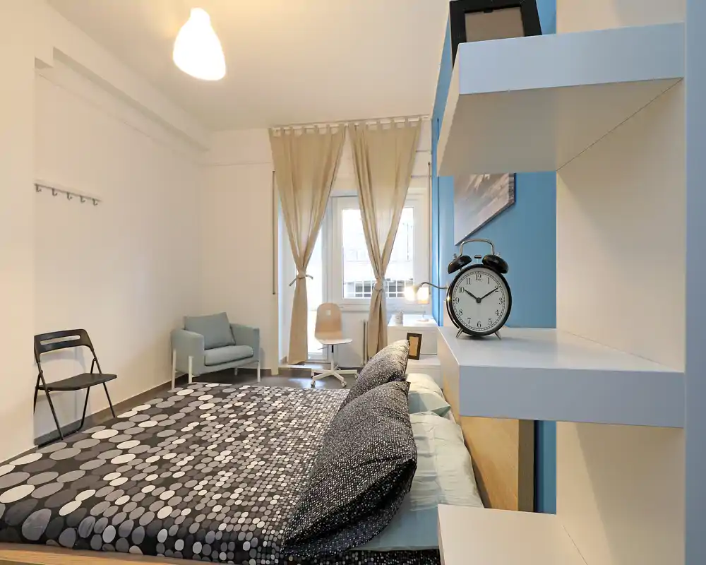 Bright, modern bedroom with a patterned double bed in the foreground, soft natural light from the window, and a cozy seating corner with an armchair and desk.
