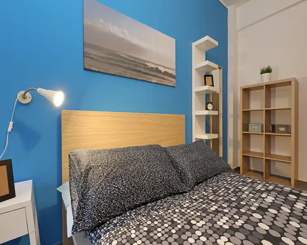 Well-staged bedroom with a patterned double bed against a vibrant blue accent wall, bedside lamp and open shelving that add functional storage and a cozy atmosphere.
