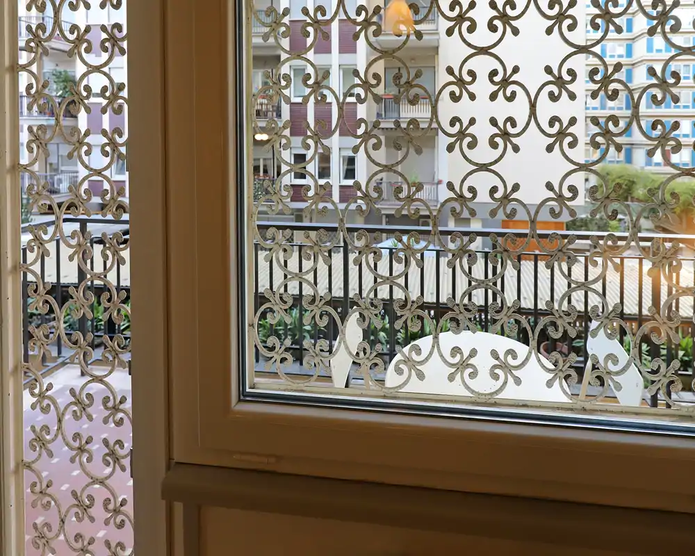 Vue depuis l'intérieur vers un petit balcon avec une grille métallique décorative et une assise partielle visible à travers la balustrade ; façades d'immeubles urbains en arrière-plan.