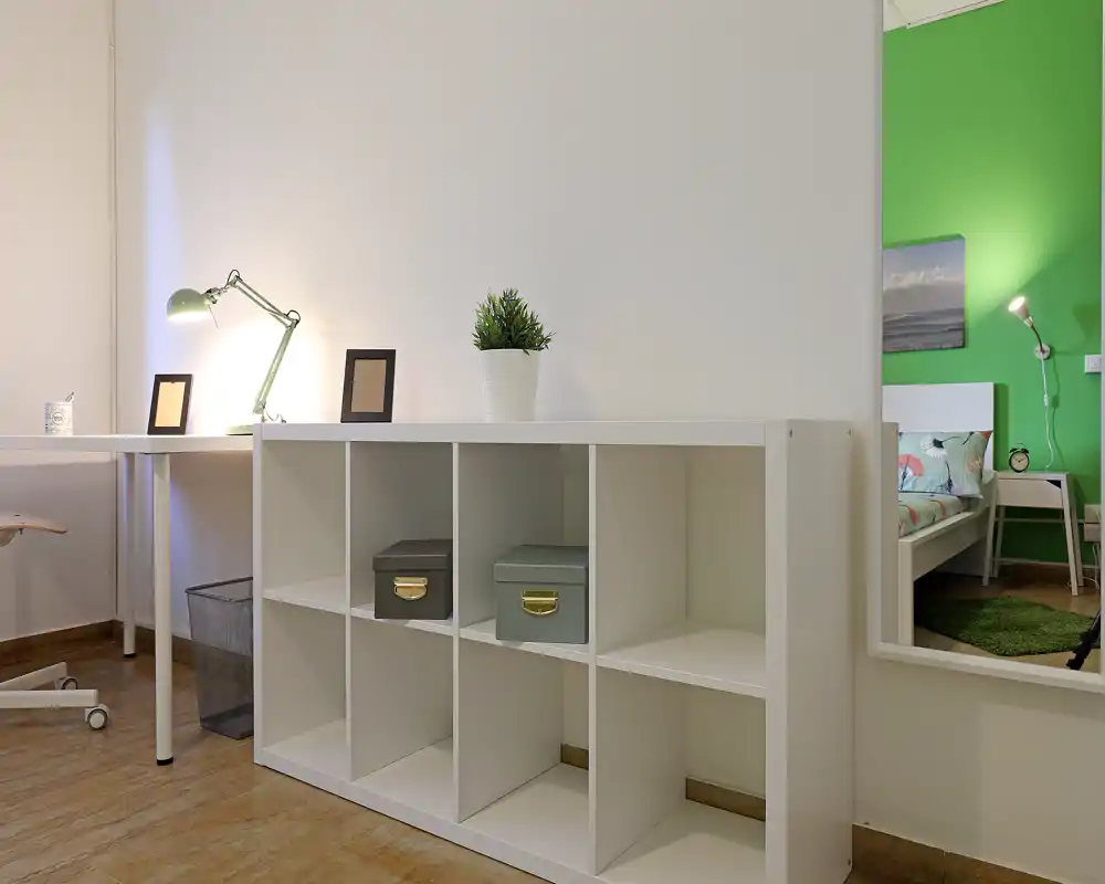 Coin de chambre moderne et lumineux vu dans le miroir : un lit blanc et une table de chevet avec lampe apparaissent en reflet, tandis que l'avant-plan montre une étagère cubique et un bureau épurés.