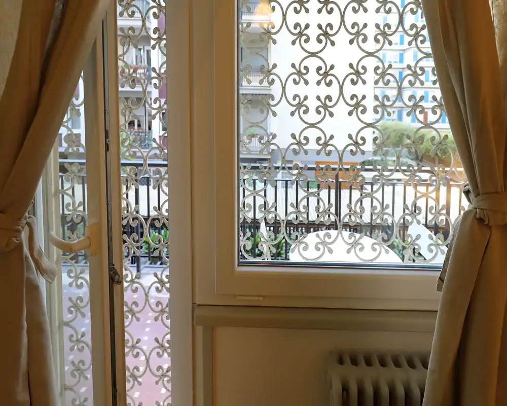 Vue à travers une fenêtre avec une grille en fer forgé décorative donnant sur un petit balcon ou une cour; des rideaux beiges encadrent la fenêtre et un radiateur est visible à l'intérieur.