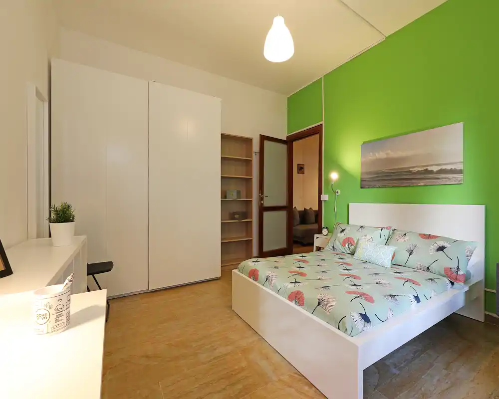 Chambre lumineuse et bien mise en scène avec lit double à motif, lampe de chevet et grand placard. Mur vert en accent et sol chaleureux, idéale pour une annonce locative.