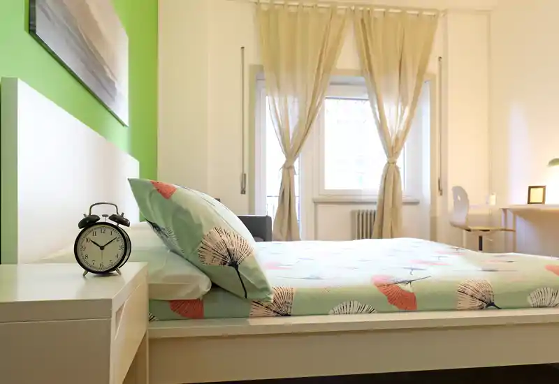 Chambre lumineuse et bien mise en scène avec une couette à motif, une table de chevet avec réveil et une fenêtre aux rideaux légers laissant passer la lumière naturelle.