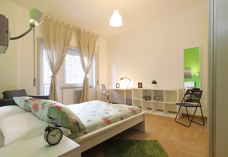 Chambre lumineuse et bien aménagée avec lit double, table de chevet et réveil, bureau et étagères, éclairée par une fenêtre aux rideaux légers.
