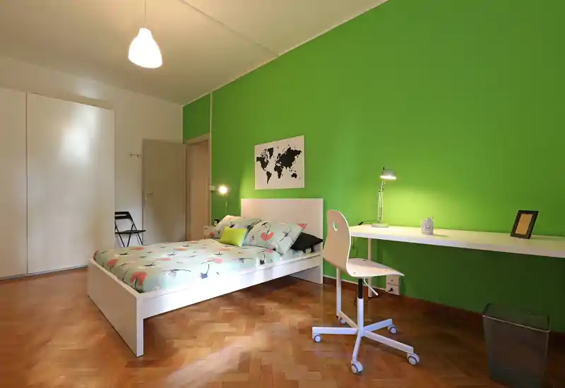 Dormitorio luminoso y bien decorado con cama doble, ropa de cama estampada y un largo escritorio frente a una pared verde intensa — ideal para estudiantes o profesionales que buscan una habitación funcional y alegre.