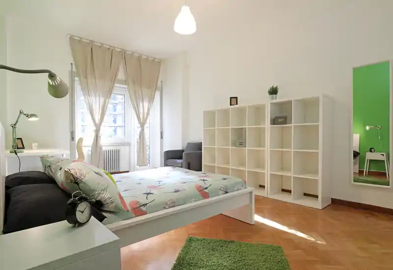 Dormitorio moderno y luminoso con cama doble en primer plano, ropa de cama estampada, alfombra verde y estantería modular. Ventana grande que aporta mucha luz natural; ambiente sencillo y bien preparado para fotos de alquiler.