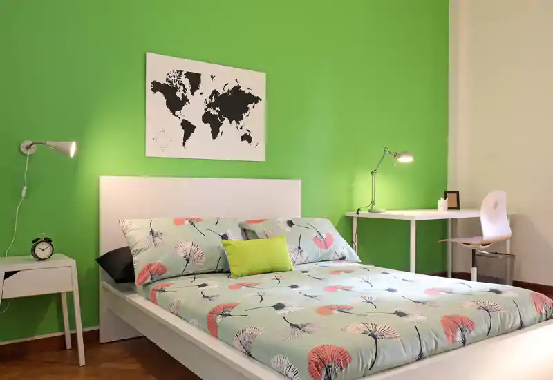 Camera da letto moderna e luminosa con un ampio letto matrimoniale, biancheria fantasia e una parete verde come accento; scrivania e comodini rendono lo spazio funzionale.