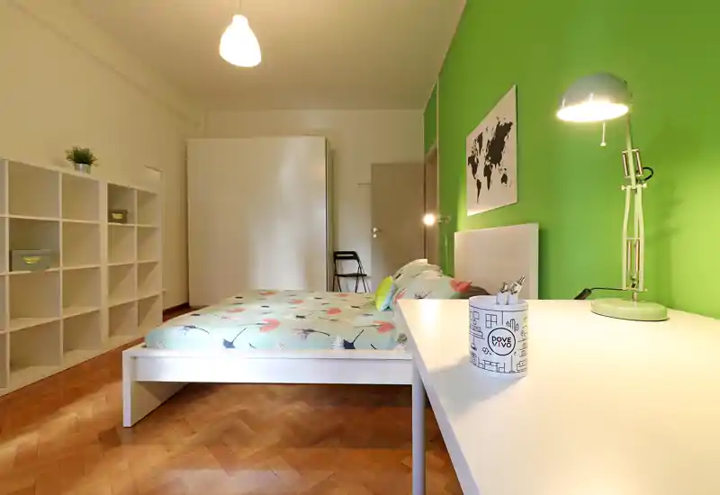 Dormitorio moderno y luminoso con cama blanca, ropa de cama estampada y pared de acento verde. La habitación incluye escritorio con lámpara, estanterías y armario, perfecta para un inquilino o pareja con estilo.