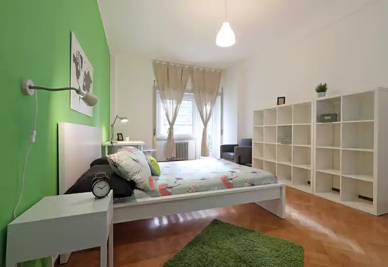 Dormitorio moderno y luminoso con cama baja blanca, ropa de cama estampada y pared verde de acento; estantería y gran ventana que aportan luminosidad y amplitud.