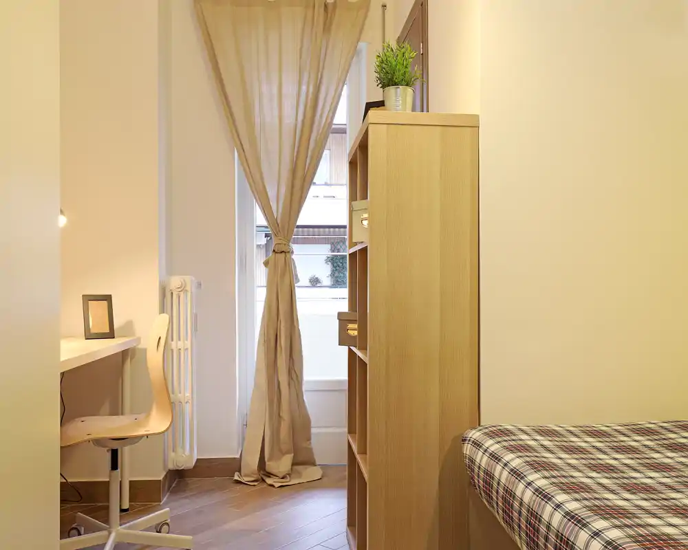 Camera da letto compatta e ben organizzata con letto singolo, libreria in legno e una scrivania vicino alla finestra che lascia entrare luce naturale morbida; toni neutri e tessuti semplici creano un'atmosfera rilassante.