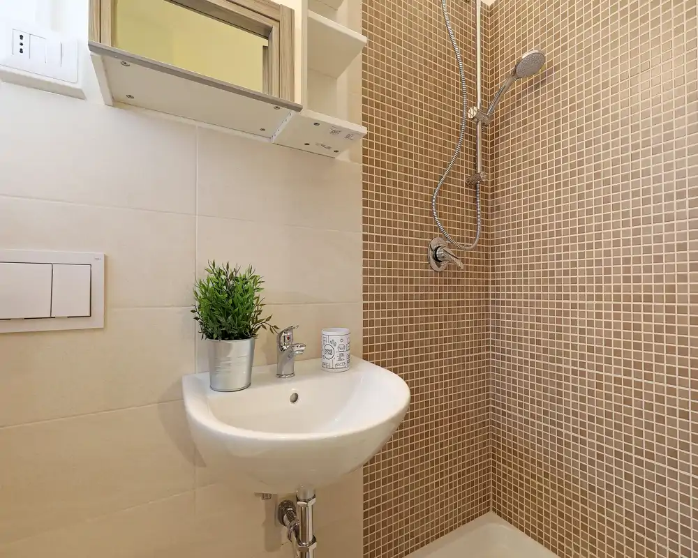 Bagno moderno e compatto con lavabo a parete, rubinetto cromato e doccia adiacente con piastrelle mosaic marroni. Finiture pulite e neutre e una piccola pianta per un tocco accogliente.