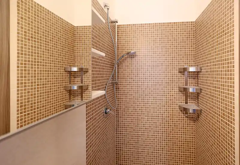 Modern shower with warm brown mosaic tiles, chrome handheld showerhead and corner shelving. Bright, well-lit stall ideal for rental listing photos.