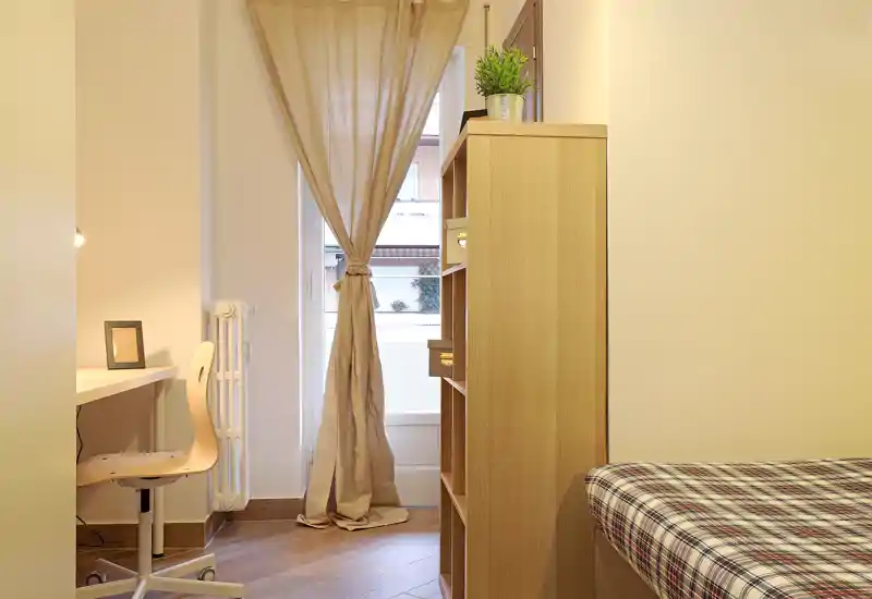 Camera da letto compatta e ben organizzata con letto singolo, libreria in legno e una scrivania vicino alla finestra che lascia entrare luce naturale morbida; toni neutri e tessuti semplici creano un'atmosfera rilassante.