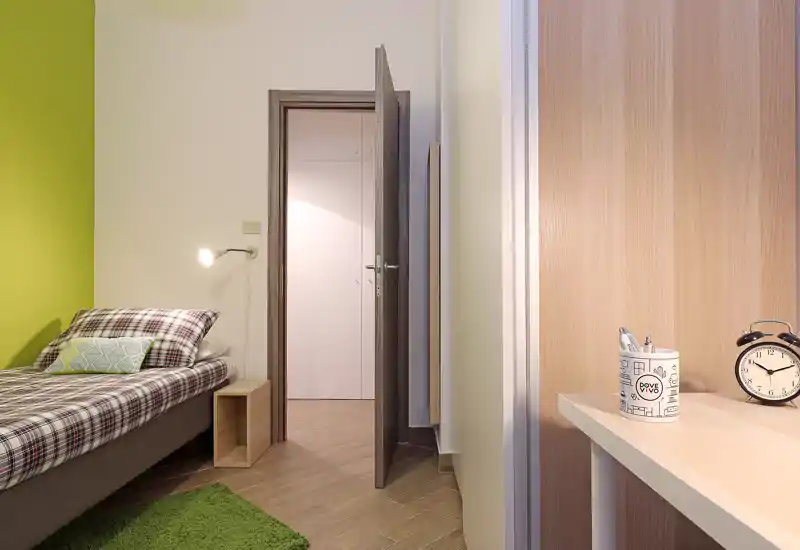 Camera singola luminosa e moderna con biancheria a quadri, lampada da lettura e un piccolo comodino in legno; pavimento in legno chiaro e parete verde a contrasto creano un’atmosfera fresca.