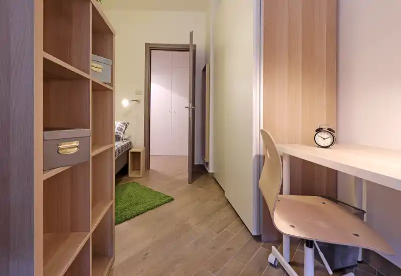 Camera da letto compatta vista da un piccolo angolo studio/ingresso: visione parziale del letto attraverso la porta aperta, scaffalatura in legno, scrivania con sedia e una sveglia, tappeto verde e pavimento in legno caldo.