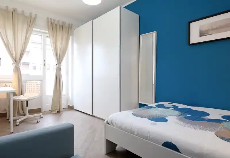Bright, modern single bedroom with a blue accent wall, white wardrobe, and patterned bedding. Natural light fills the room from a window with light curtains, and a small desk and chair make it suitable for study.