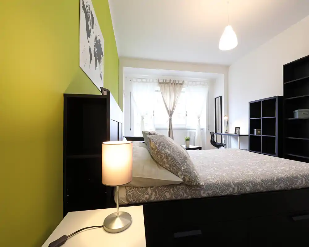 Chambre moderne et lumineuse avec literie à motif, lampe de chevet et grande fenêtre apportant une belle lumière naturelle. Mur accent vert, rangements et coin bureau pour un séjour confortable.