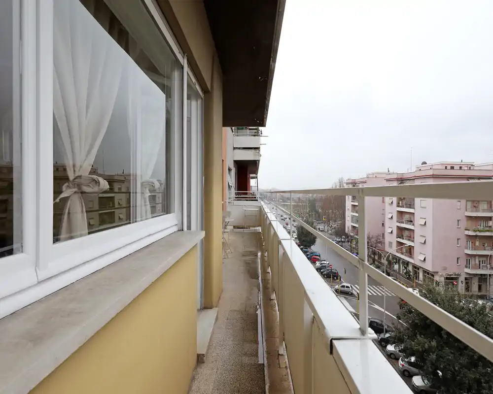 Balcon étroit et long donnant sur une rue résidentielle avec une rambarde métallique et des immeubles voisins ; lumière naturelle grise et sol carrelé.