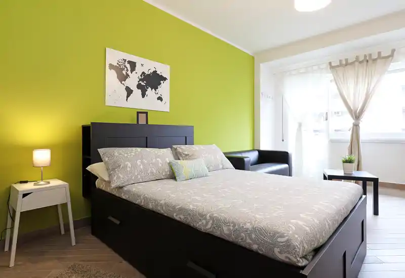 Chambre lumineuse et bien mise en scène avec lit double devant un mur vert, linge de lit doux, lampe de chevet et lumière naturelle provenant d'une grande fenêtre.