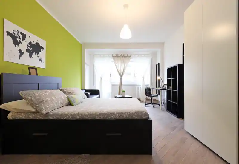 Chambre moderne et lumineuse avec grand lit double, mur d'accent vert, rangements sous le lit et coin bureau près de la fenêtre ; décoration soignée, idéale pour locations.