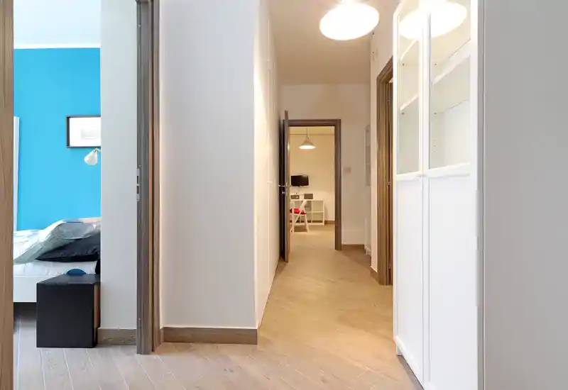 View of a hallway with a partially visible bedroom on the left: you can see the bed frame, pillows and bedside table through the doorway. Bright, modern corridor with wood flooring and built-in storage.