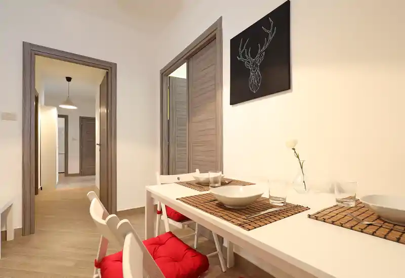 Bright, modern dining corner with a white table set for four, woven placemats and red seat cushions; minimalist wall art and a small vase complete the inviting space.