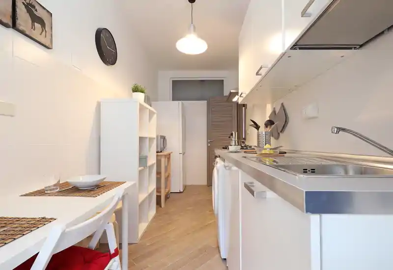 Bright, compact galley kitchen with white cabinets, stainless sink and small dining area — clean modern styling and warm wood flooring.