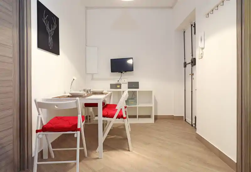 Compact dining area with a small white table, folding chairs with red cushions and a minimal shelving unit. Well-lit, tidy and styled for two diners.