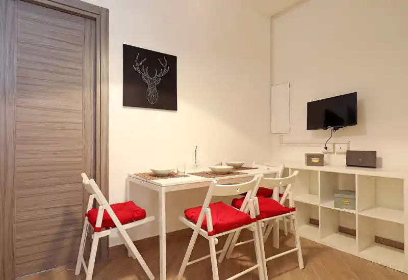 Compact dining corner with a small white table and four folding chairs dressed with red cushions; simple table setting and shelving unit create a cozy, modern mealtime spot.