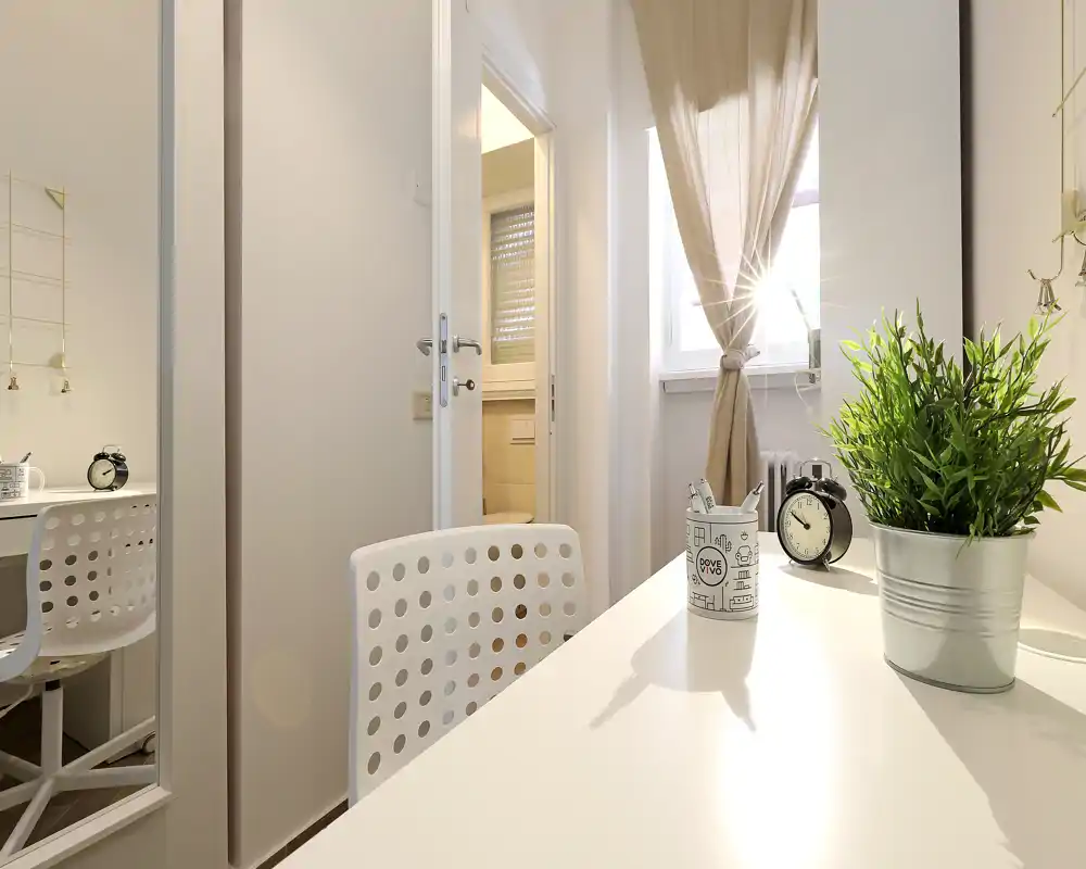 Bright, minimal workspace corner with a white desk, perforated chair, decorative plant and an alarm clock by a sunlit window — a cozy nook suitable for reading or working.