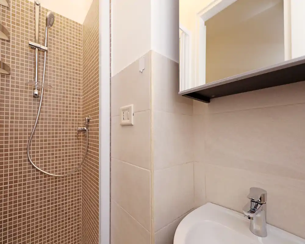 Compact bathroom featuring a tiled shower with handheld mixer and a small sink with modern faucet. Neutral beige tiles create a clean, minimal look.