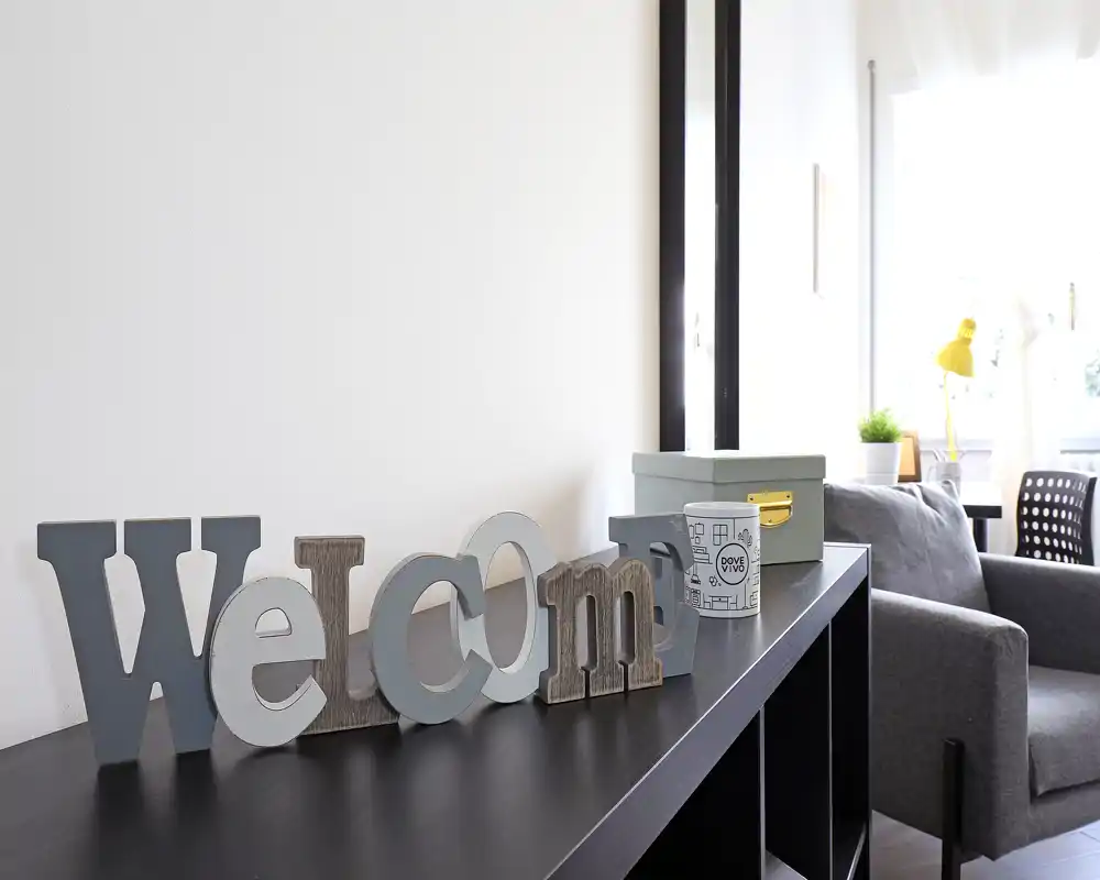 Espace de vie lumineux et moderne avec un fauteuil gris, des lettres décoratives « welcome » sur un meuble et la lumière naturelle d'une fenêtre — un coin cosy parfait pour accueillir.