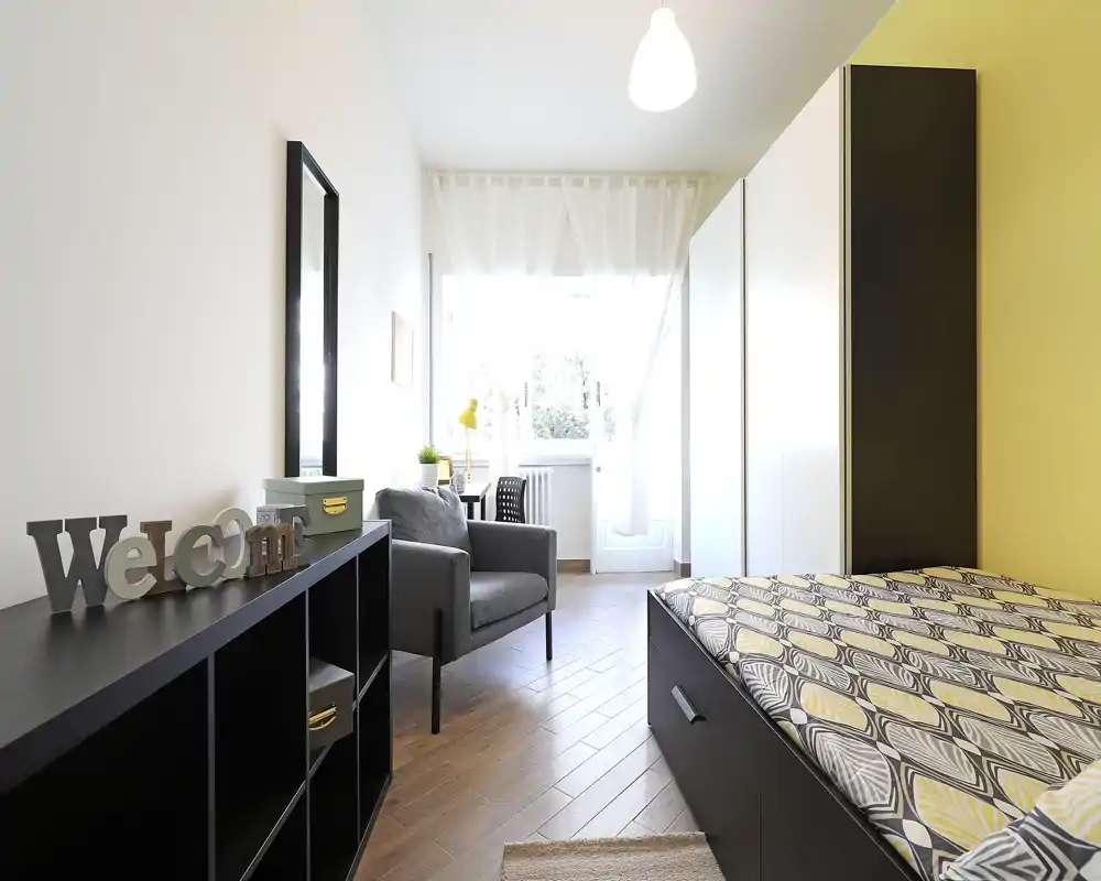 Chambre moderne et lumineuse avec une couette à motifs, un fauteuil confortable et une armoire près d'une fenêtre ensoleillée — idéale pour un séjour cosy.