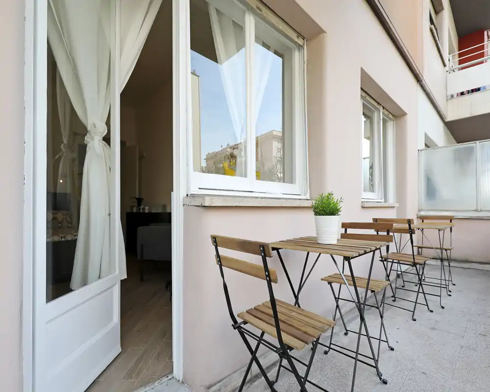 Balcon ensoleillé avec petites tables et chaises bistrot en bois, sol carrelé et accès direct depuis l'appartement par une porte vitrée — parfait pour le café du matin.