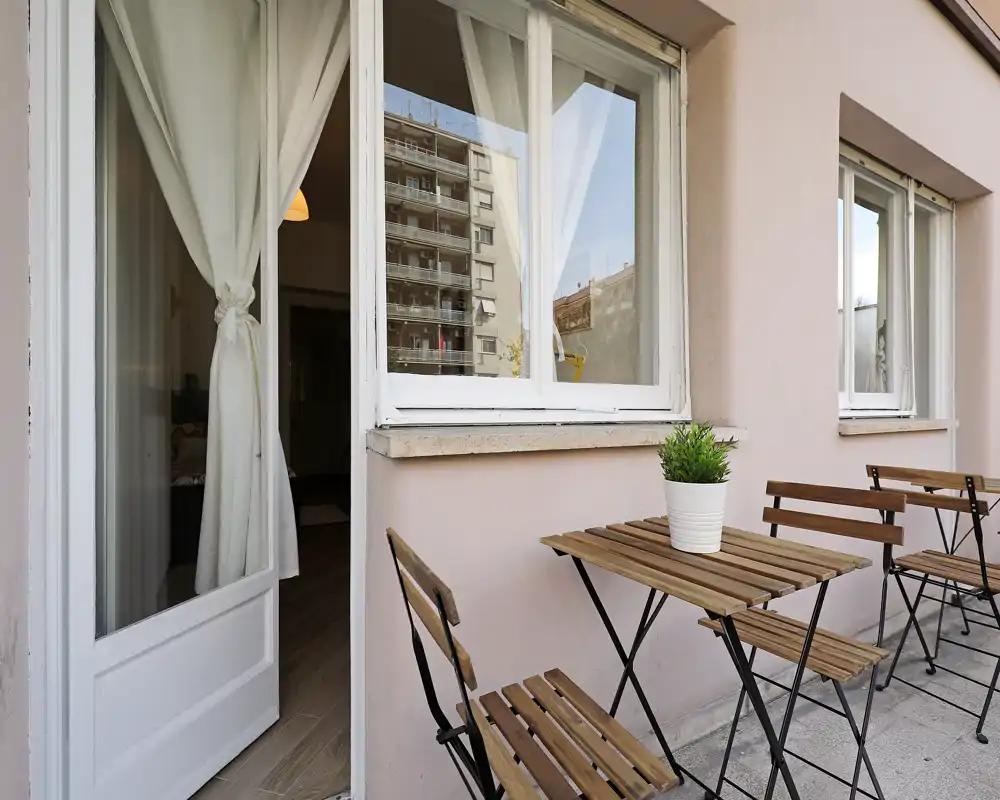 Petit balcon meublé avec table et chaises pliantes en bois, une plante en pot sur la table et vue sur l’appartement à travers une porte et des fenêtres aux encadrements blancs. Idéal pour un café matinal dans un espace urbain compact.