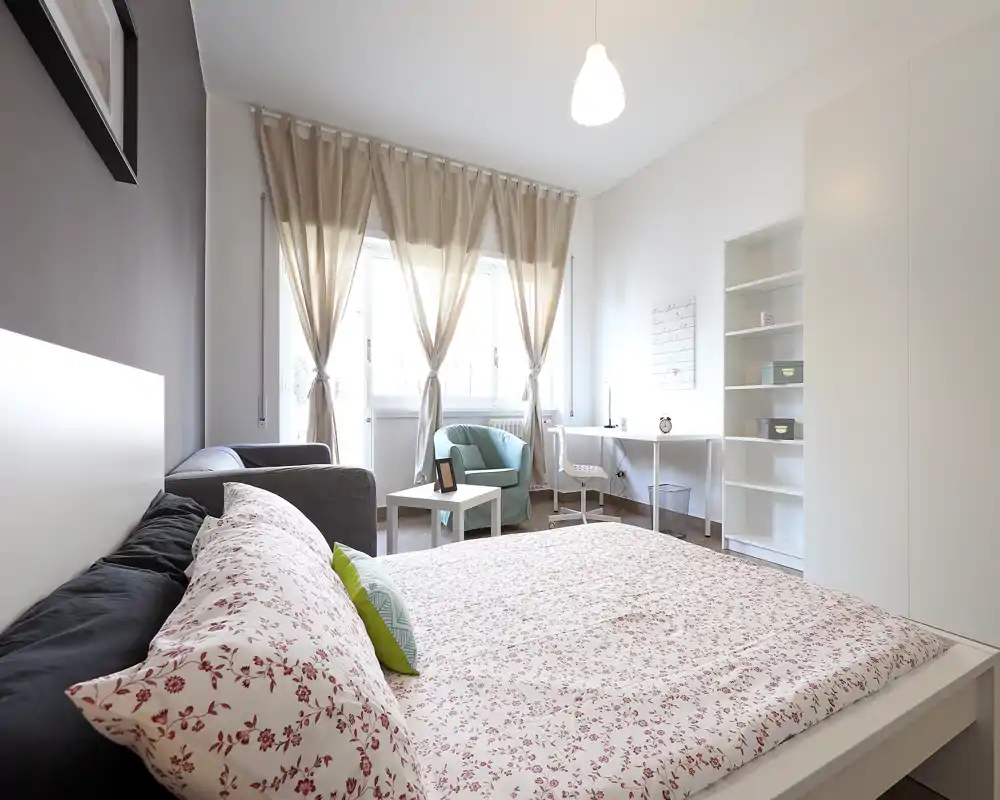 Dormitorio moderno y luminoso con cama doble en primer plano, edredón estampado y cojines, un rincón de lectura junto a la ventana y un pequeño escritorio — ideal para estancias cómodas o alquileres de larga duración.