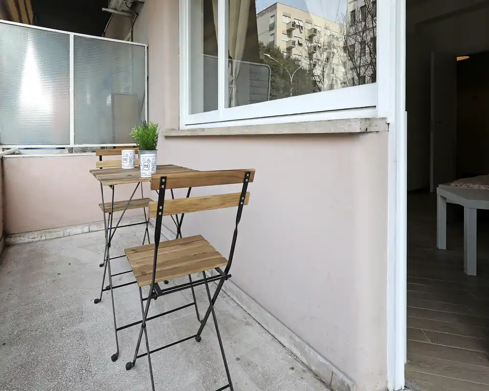 Pequeño balcón amueblado con mesa bistró de madera y dos sillas plegables, una planta en maceta y una pantalla de privacidad — ideal para el café de la mañana.