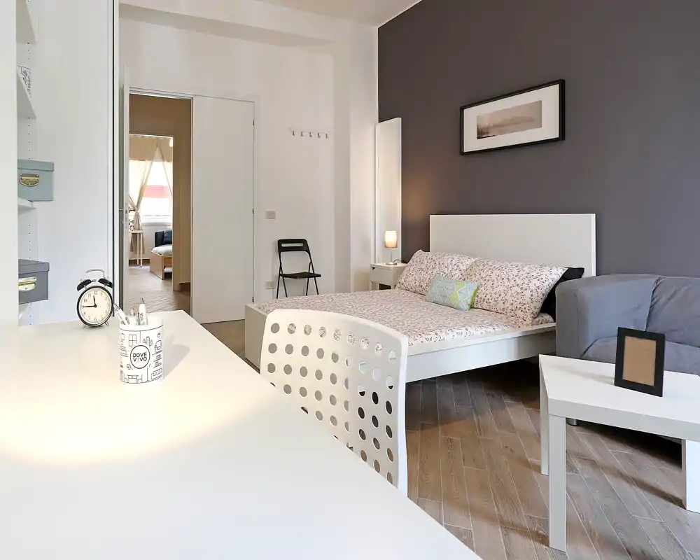 Habitación luminosa y moderna con cama blanca, ropa de cama estampada y lámpara de noche acogedora; suelo de madera y paleta neutra y estilosa.