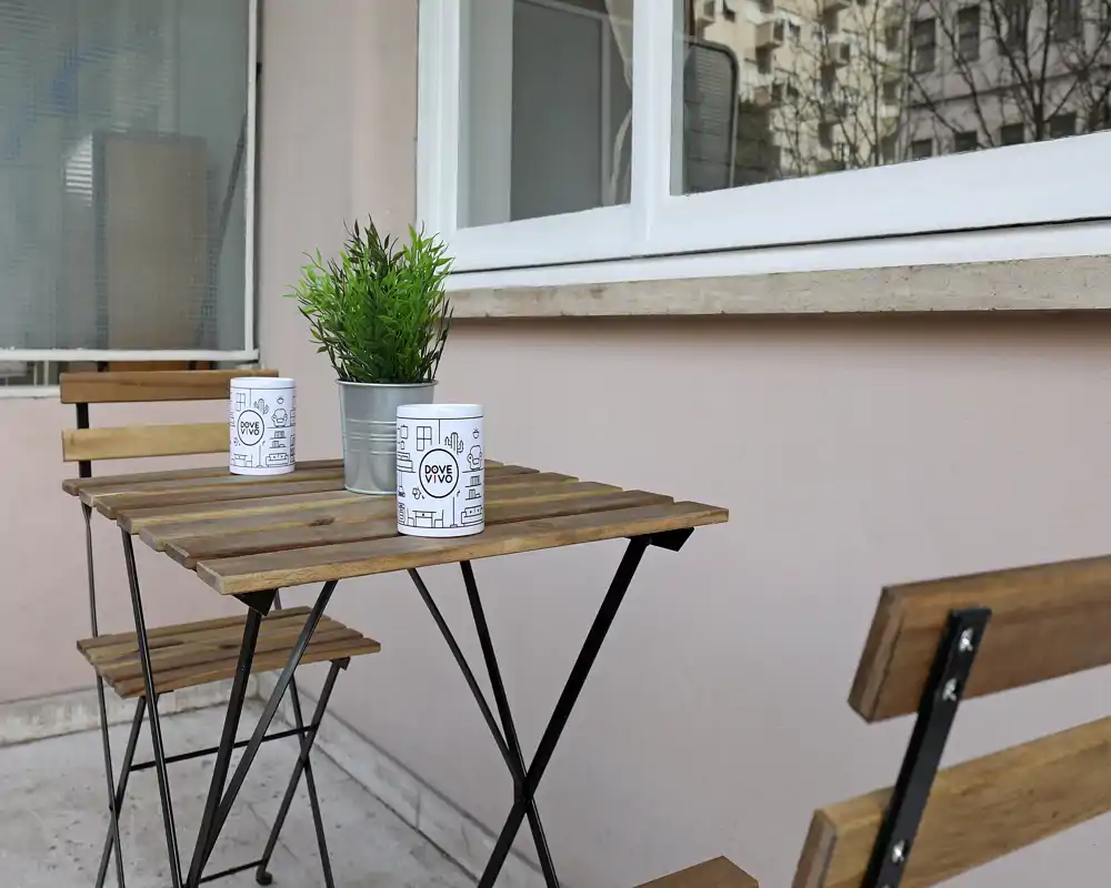 Pequeño balcón amueblado con mesa bistró de madera, dos sillas plegables y una planta decorativa — un rincón acogedor para el café.