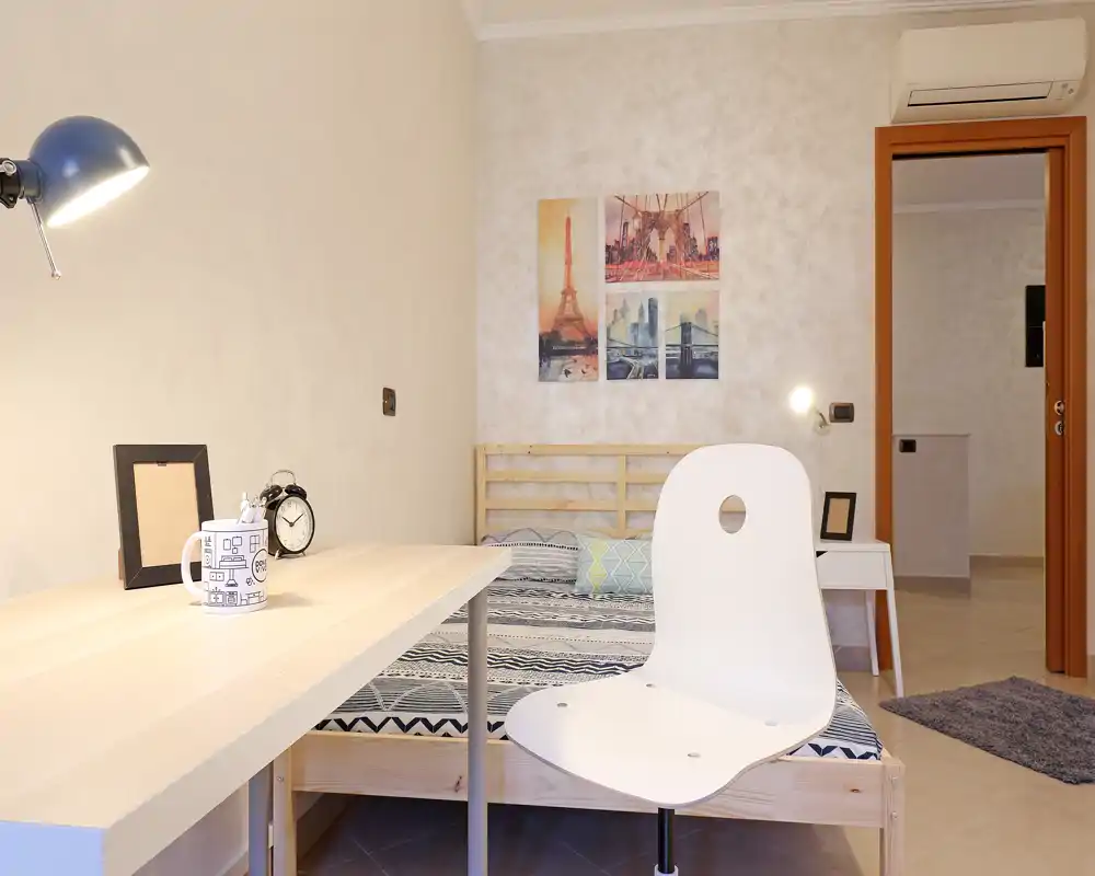 Dormitorio luminoso y ordenado con una cama de madera sencilla, ropa de cama con estampado y un escritorio en primer plano. El arte en la pared y la iluminación suave generan un espacio acogedor y funcional, ideal para una persona.