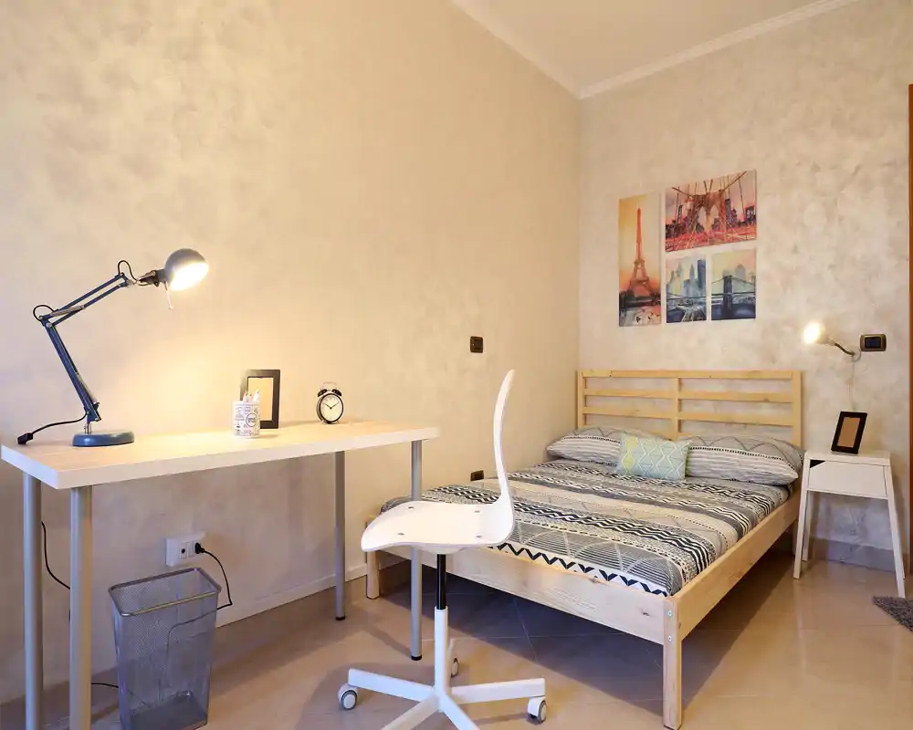 Dormitorio luminoso y ordenado con cama doble de madera, ropa de cama estampada, escritorio y lámpara de trabajo, ideal para descansar y estudiar.