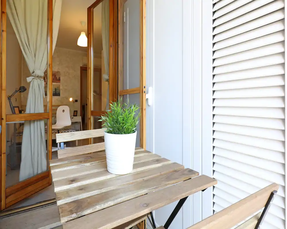 Pequeño balcón amueblado con mesa de madera y planta en maceta, que se abre a una habitación interior mediante puertas de cristal. Luminoso, limpio y acogedor, ideal para tomar café por la mañana.
