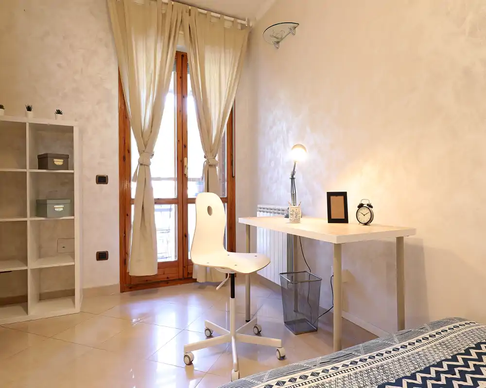 Rincón de dormitorio luminoso y amueblado con sencillez, con un pequeño escritorio, silla y vista parcial de la cama. Cortinas claras cubren la puerta balcón dejando entrar abundante luz natural.