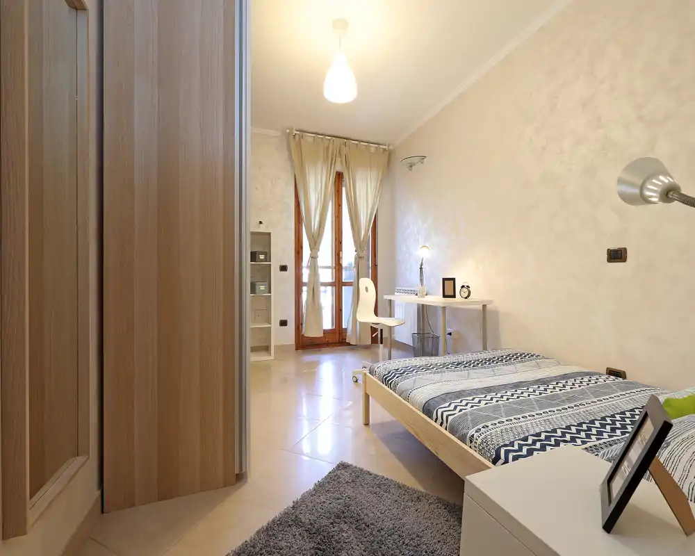 Dormitorio moderno y luminoso con cama baja de madera, ropa de cama estampada, escritorio junto al balcón y luz cálida — ideal para una estancia cómoda.