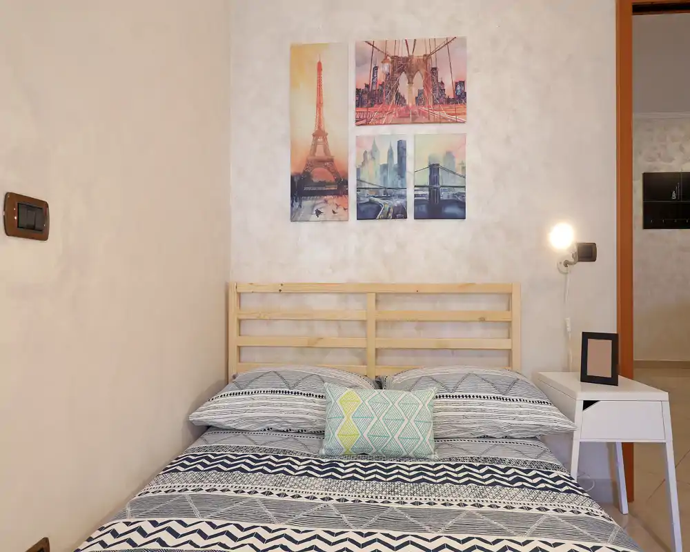 Dormitorio luminoso y ordenado con ropa de cama de estampado, cabecero de madera con lamas y cuadros urbanos encima; mesita con lámpara que aporta calidez.