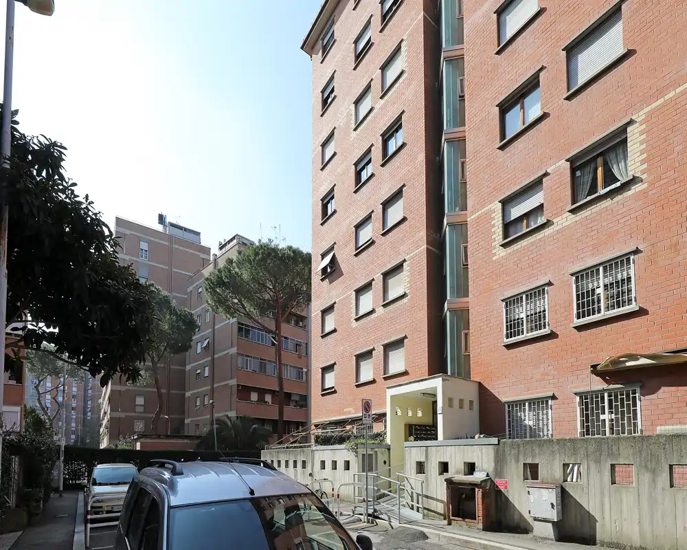 Vista exterior de un edificio de apartamentos con entrada a nivel de calle, coches aparcados y árboles alrededor con luz diurna.